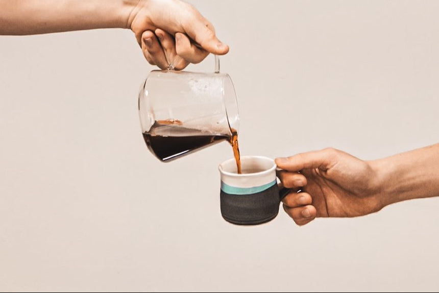 Two hands exchanging a glass carafe of coffee for a small cup with a beige background